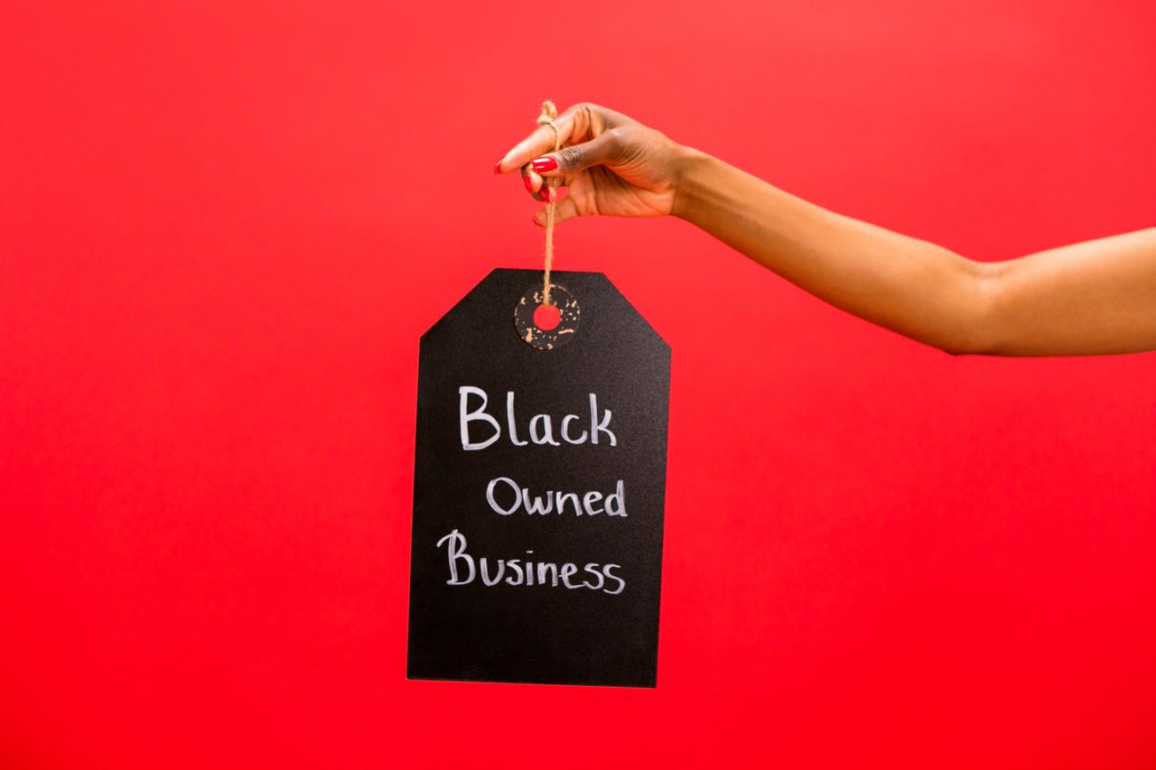 A hand holding a Black Owned Business sign against a vibrant red background, symbolizing support and diversity.